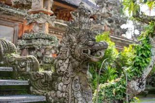 Ilustrasi: Patung naga batu berukir khas Bali di tangga pura dengan latar pepohonan hijau.