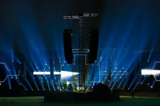 Ilustrasi: Panggung konser luar ruangan pada malam hari dengan sorotan lampu biru dan pengeras suara gantung.