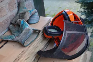 Ilustrasi: Helm keselamatan oranye dengan penutup telinga dan sarung tangan kerja di atas papan kayu.