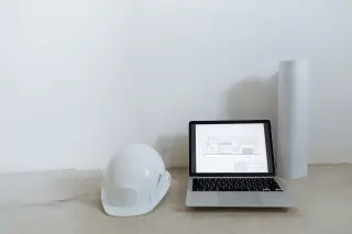 Ilustrasi: Helm proyek putih, laptop menampilkan denah bangunan, dan gulungan kertas di lantai.