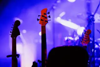 Ilustrasi: Deretan kepala gitar dan bas di atas panggung dengan sorotan lampu konser berwarna ungu kebiruan.