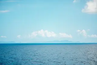 Ilustrasi: Pemandangan laut biru luas dengan garis cakrawala dan pulau di kejauhan di bawah langit cerah.