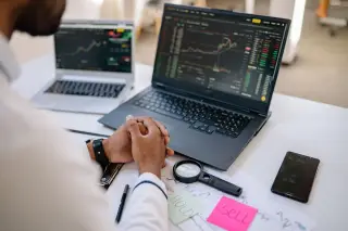 Ilustrasi: Seseorang duduk di depan laptop yang menampilkan grafik pergerakan harga saham di meja kerja.