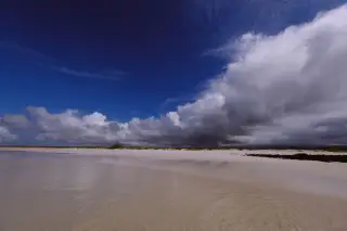 Ilustrasi: Pemandangan pantai berpasir putih luas di bawah langit biru berawan tebal.