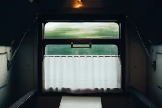 Ilustrasi: Interior kompartemen kereta dengan jendela bertirai putih menghadap pemandangan hijau yang bergerak.