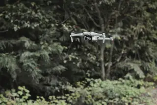Ilustrasi: Sebuah drone berwarna abu-abu terbang di depan deretan pepohonan rindang.