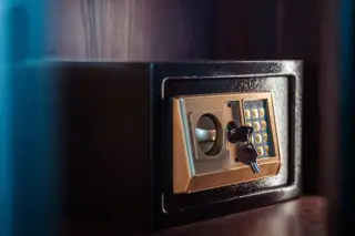 Ilustrasi: Sebuah brankas elektronik kecil warna hitam dengan kunci terpasang di atas lemari kayu.