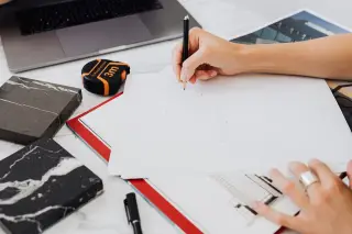Ilustrasi: Tangan seseorang sedang membuat sketsa arsitektur dengan pensil di meja kerja bersama meteran dan laptop.