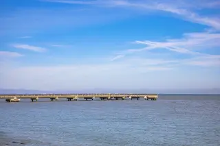 Ilustrasi: Dermaga panjang yang sepi membentang di atas laut tenang di bawah langit biru cerah.