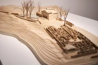 Ilustrasi: Maket arsitektur kayu yang menampilkan kontur tanah, bangunan, dan pepohonan miniatur di atas meja.