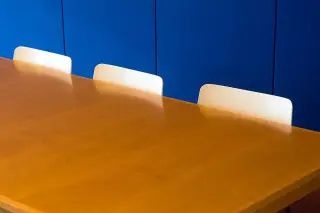 Ilustrasi: Meja kayu panjang dengan deretan sandaran kursi putih berlatar dinding biru.