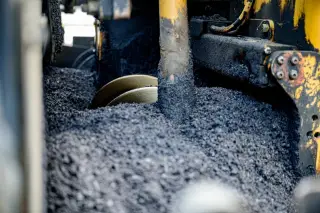 Ilustrasi: Tumpukan material aspal hitam di dalam mesin pengaspal jalan berwarna kuning.