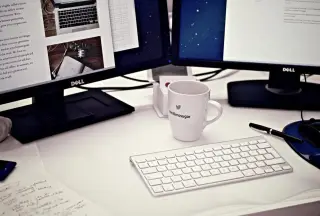 Ilustrasi: Meja kerja kantor dengan dua monitor komputer, papan ketik, dan sebuah cangkir.