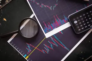 Ilustrasi: Kaca pembesar, ponsel pintar, dan cetakan grafik pergerakan saham di atas meja gelap.