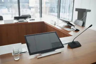 Ilustrasi: Meja rapat kayu dengan layar monitor kecil, mikrofon konferensi, dan gelas kosong.