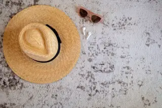 Ilustrasi: Topi jerami, kacamata hitam, dan earphone nirkabel di atas karpet bertekstur.