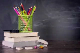 Ilustrasi: Tumpukan buku dan wadah pensil warna di atas meja dengan latar papan tulis hitam.