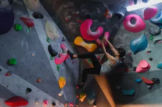 Ilustrasi: Seorang wanita sedang memanjat dinding bouldering dalam ruangan dengan berbagai pegangan warna-warni.