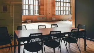 Ilustrasi: Meja rapat kayu panjang dengan deretan kursi hitam kosong di dalam ruangan.