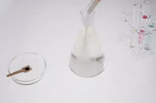 Ilustrasi: Peralatan laboratorium kaca berupa labu erlenmeyer, cawan petri, dan tabung reaksi di atas meja putih.