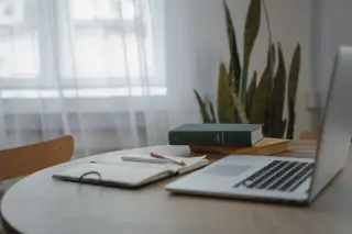 Ilustrasi: Sebuah laptop, buku catatan terbuka, dan tumpukan buku di atas meja kayu dekat jendela.