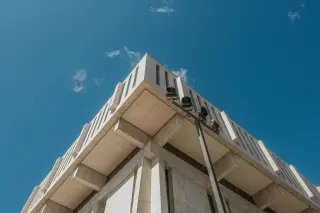Ilustrasi: Sudut atas gedung perkantoran bertingkat dengan fasad beton berlatar langit biru.