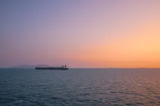 Ilustrasi: Kapal kargo besar berlayar di laut lepas berlatar langit senja berwarna jingga dan ungu.