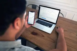 Ilustrasi: Seseorang melihat grafik pergerakan saham melalui ponsel dan laptop di meja kayu.