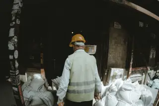 Ilustrasi: Seorang pekerja berompi keselamatan dan helm proyek kuning tampak dari belakang dekat karung-karung.