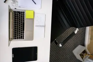 Ilustrasi: Pemandangan dari atas meja kerja putih dengan laptop, tablet, catatan, dan kursi kantor hitam.