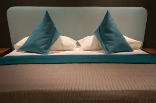 Ilustrasi: Tempat tidur hotel modern dengan bantal biru kehijauan dan sandaran kepala senada.