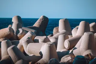Ilustrasi: Tumpukan beton tetrapod pemecah ombak di tepi laut biru dengan latar langit cerah.