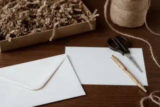 Ilustrasi: Amplop putih, kartu kosong, pena kayu, dan gunting di atas meja kayu.