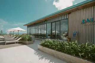 Ilustrasi: Bangunan resor modern berpintu kaca dengan teras luar ruangan, kursi santai, dan payung pantai.