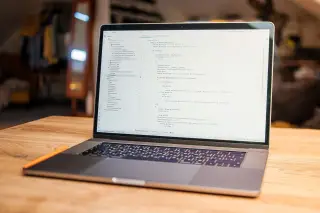 Ilustrasi: Sebuah laptop menyala menampilkan baris kode di atas meja kayu dengan latar belakang ruangan.