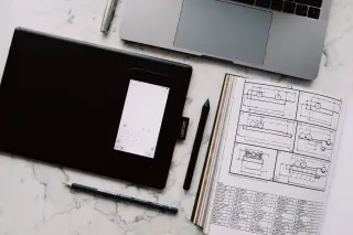 Ilustrasi: Tablet grafis, ponsel pintar, laptop, dan buku sketsa teknis di atas meja marmer.