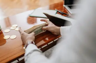 Ilustrasi: Seseorang sedang memegang segepok uang kertas dolar AS di atas meja kayu.