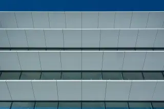 Ilustrasi: Tampilan simetris fasad gedung modern dengan panel putih dan kaca di bawah langit biru cerah.
