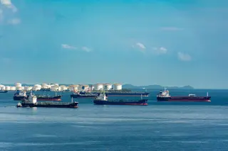 Ilustrasi: Kapal-kapal tanker berlayar di laut dengan tangki minyak di tepi pantai di kejauhan.