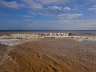 Ilustrasi: Ombak air laut yang keruh dan kecokelatan menerjang pesisir pantai di bawah langit biru berawan.