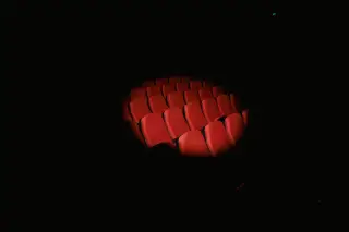 Ilustrasi: Deretan kursi teater berwarna merah yang disorot cahaya di tengah ruangan gelap.