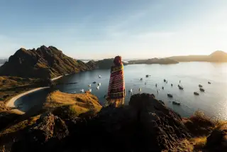 Ilustrasi: Seseorang berselimut kain etnik berdiri di bukit memandang teluk laut dengan banyak kapal wisata.
