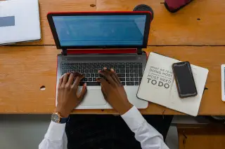 Ilustrasi: Seseorang mengetik pada laptop di atas meja kayu di samping buku catatan dan ponsel.