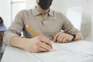 Ilustrasi: Seseorang bermasker duduk di meja kelas sedang mengisi lembar ujian dengan pensil.