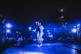Ilustrasi: Seorang penampil di atas panggung konser terbuka menghadap penonton di malam hari di bawah lampu biru.