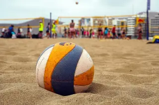 Ilustrasi: Bola voli di atas pasir pantai dengan latar belakang net dan pemain yang buram.