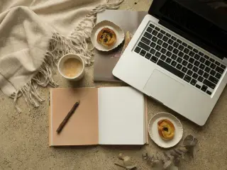 Ilustrasi: Laptop, buku catatan terbuka, kopi, dan kue tertata rapi di atas meja.