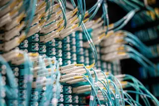 Ilustrasi: Deretan kabel serat optik berwarna biru toska terpasang pada panel server jaringan.