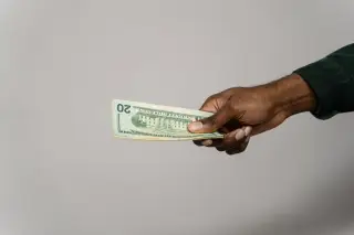 Ilustrasi: Sebuah tangan memegang beberapa lembar uang kertas dua puluh dolar AS dengan latar belakang abu-abu polos.