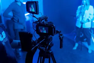 Ilustrasi: Kamera video profesional merekam seseorang di studio dengan pencahayaan bernuansa biru.